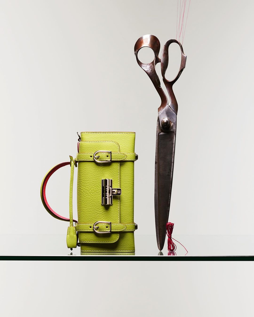 Studio shot of a lime-green leather handbag with buckles and large, antique-style shears, both resting on a reflective surface against a neutral background.