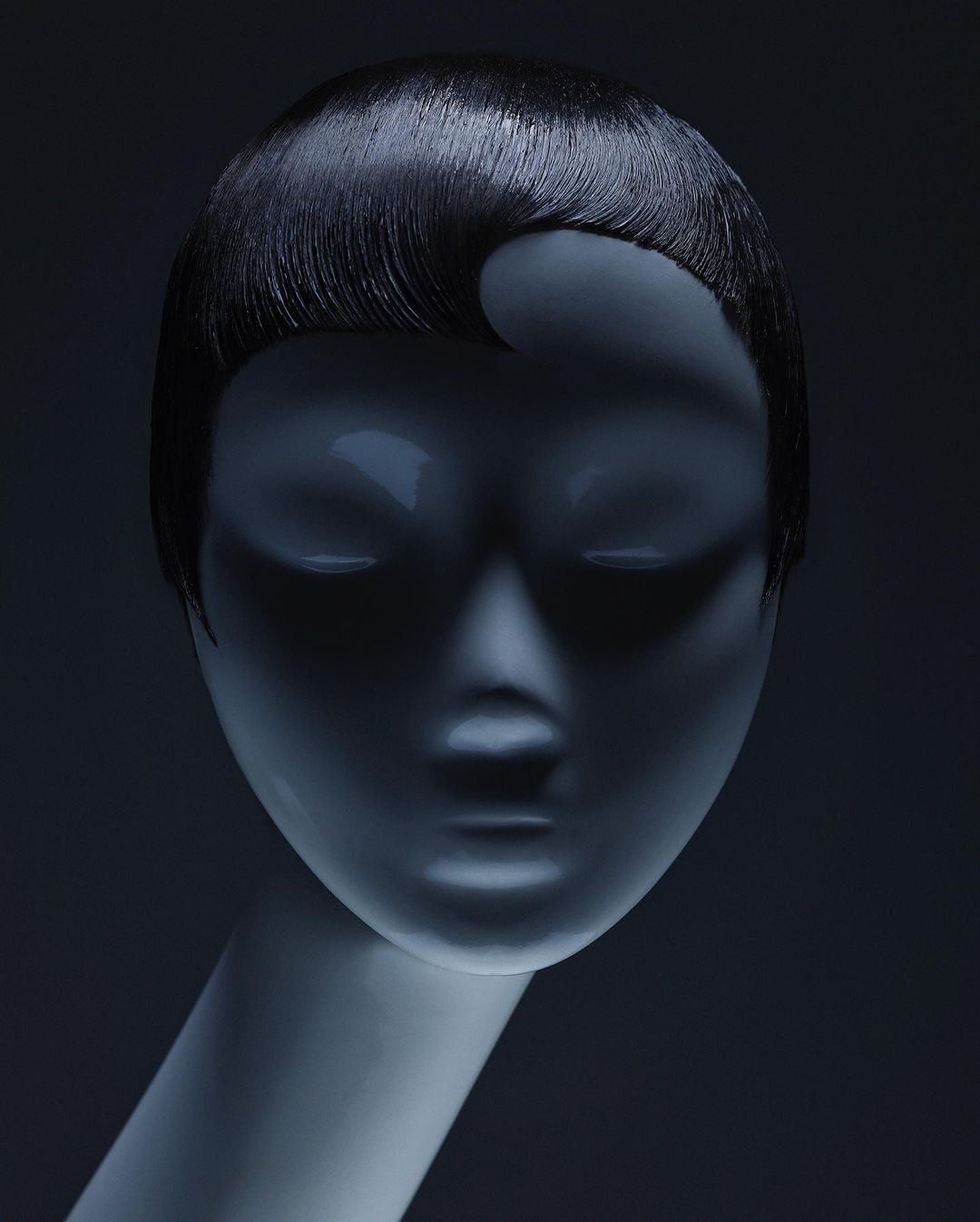 Close-up of a mannequin head with a short, dark, wavy hairstyle, displayed against a dark gray background in a studio-lit setting.