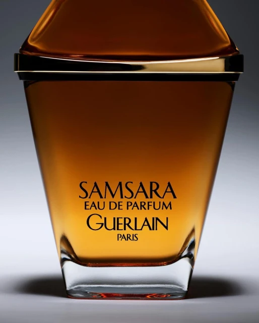 Close-up studio shot of a golden-brown glass Guerlain Samsara Eau de Parfum bottle with black text against a graduated gray background.