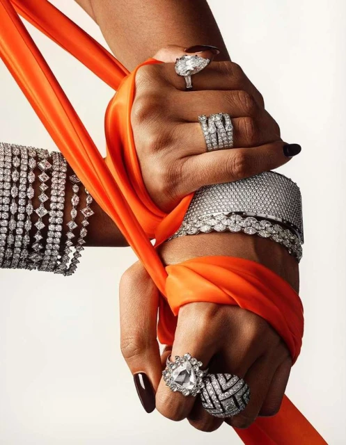Close-up of two arms adorned with diamond bracelets and rings, clutching a bright orange fabric against a neutral background.