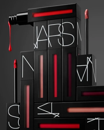 Cosmetics display: NARS brand lipsticks and applicators in black packaging, arranged in a staggered geometric stack against gray background.