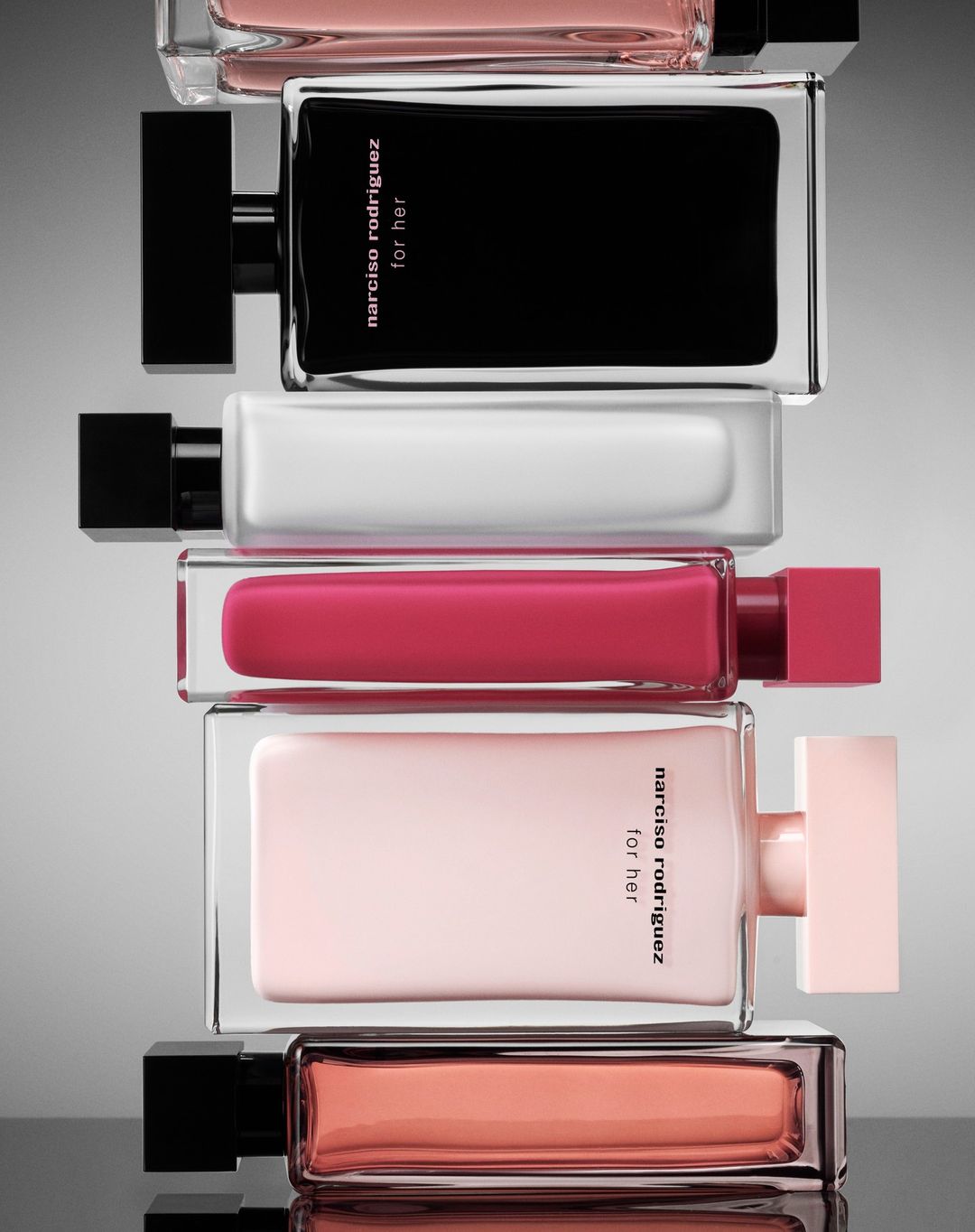 Stacked arrangement of Narciso Rodriguez For Her perfume bottles in various colors including black, white, pink, and peach.