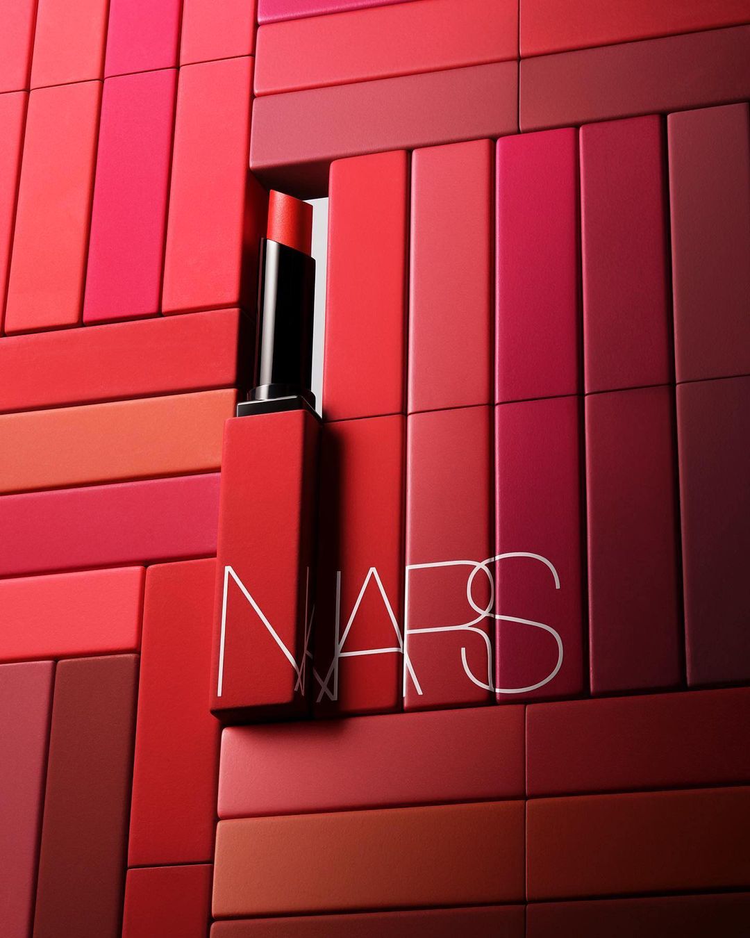 NARS lipstick tube and lettering against blocks in shades of red, coral, and pink, arranged in a flat lay style.