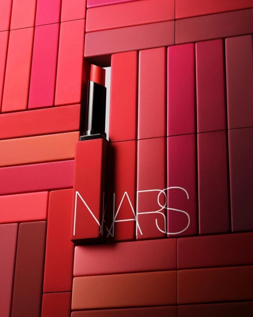 NARS lipstick tube and lettering against blocks in shades of red, coral, and pink, arranged in a flat lay style.