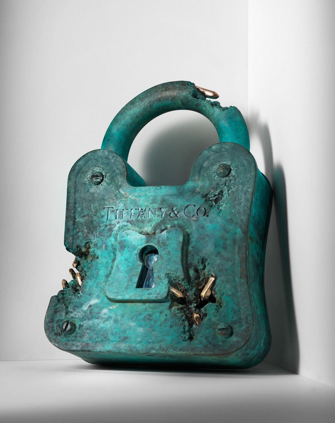 Distressed teal Tiffany & Co. padlock sculpture with gold accents positioned on a white surface against a gradient white background.
