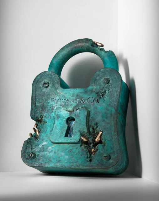 Distressed teal Tiffany & Co. padlock sculpture with gold accents positioned on a white surface against a gradient white background.