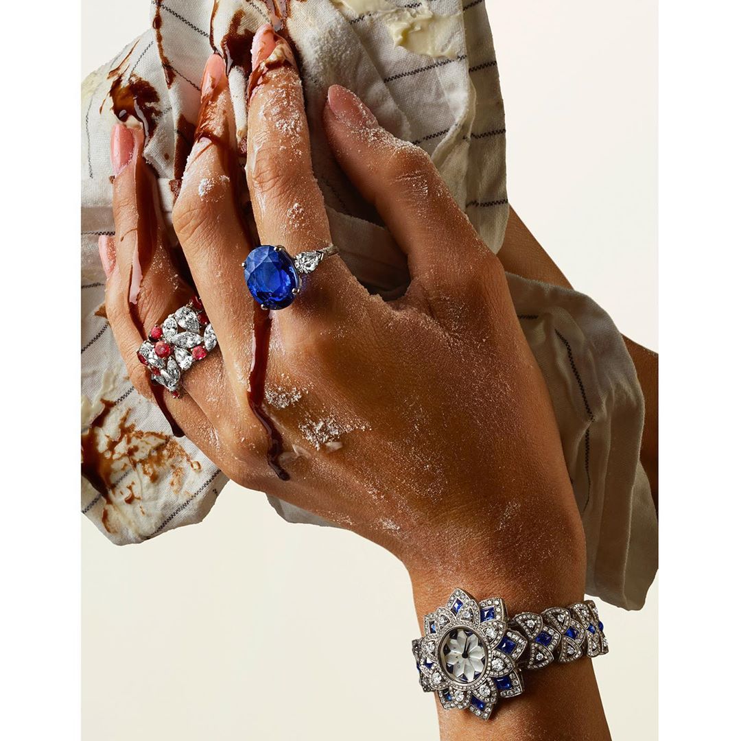 Jewellery advertisement featuring a person's hand adorned with blue sapphire rings and a diamond watch, all dusted with flour.