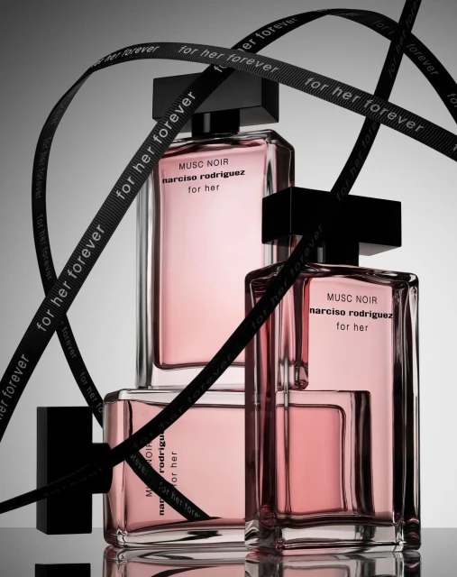 Two Narciso Rodriguez Musc Noir perfume bottles with black caps and a ribbon with repeating text on a reflective gray surface.