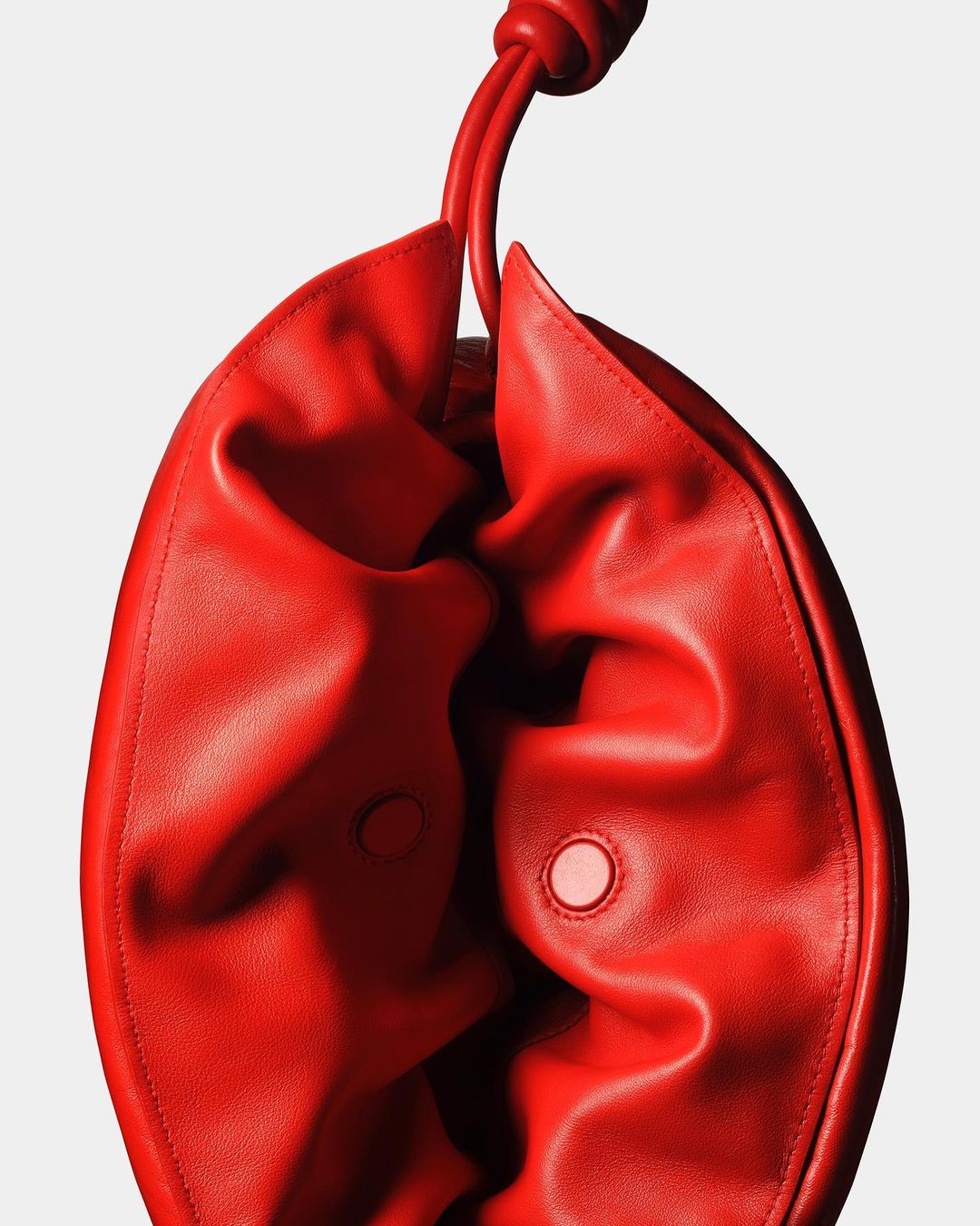 Close-up of a bright red leather shoulder bag with a knotted handle. The bag has two buttons and is photographed against a white background.