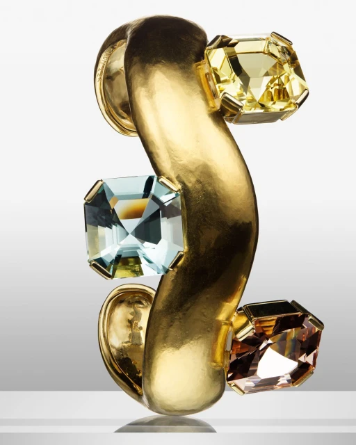 Close-up of a gold-toned metal cuff bracelet featuring three square gemstones in yellow, brown, and blue against a neutral gradient background.