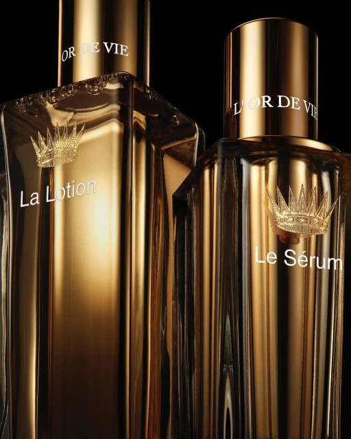 Two glass bottles of L'Or de Vie skincare products, one labeled 'La Lotion' and the other 'Le Sérum', stand against a dark backdrop.