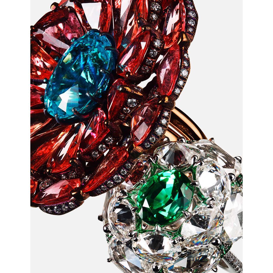 Two flower-shaped rings featuring red, turquoise, clear, and green gems against a white background in a jewellery close-up shot.