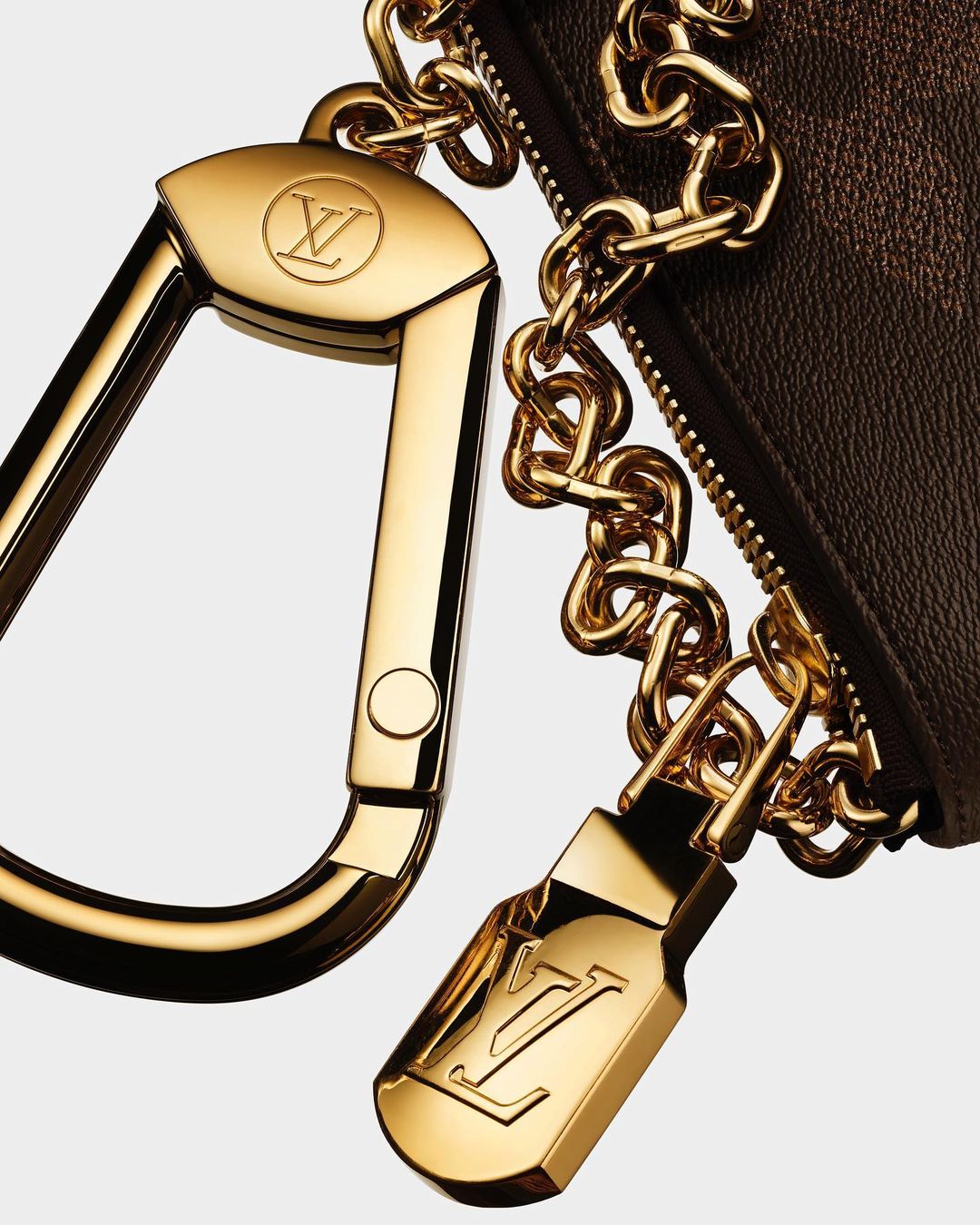 Close-up of a gold Louis Vuitton bag accessory with a carabiner clip, chain, and branded zipper pull, set against a neutral background.