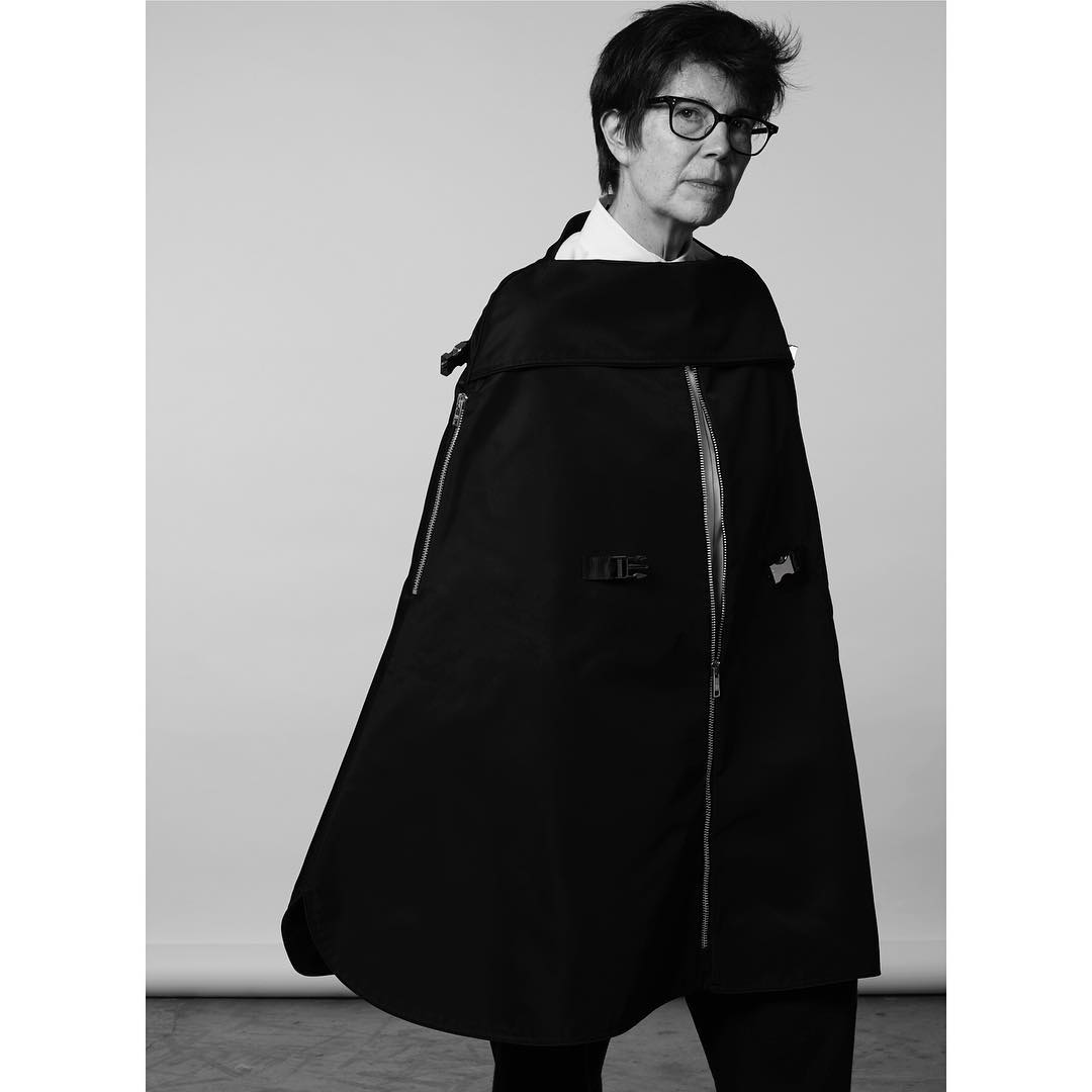 Black and white portrait of an adult female wearing glasses and a black cape with silver zippers, standing in a neutral-toned studio.