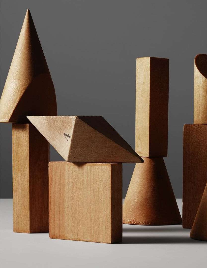 Geometric solids in wood: cones, cubes, pyramids. A collection of vintage wooden 3D geometric shapes on a white surface against a gray backdrop.