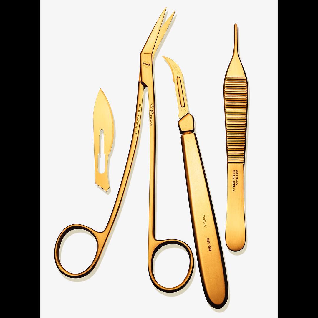 Gold-tone medical instruments, including surgical scissors, scalpel blades, and a tweezer, are arranged on a clean, white background.