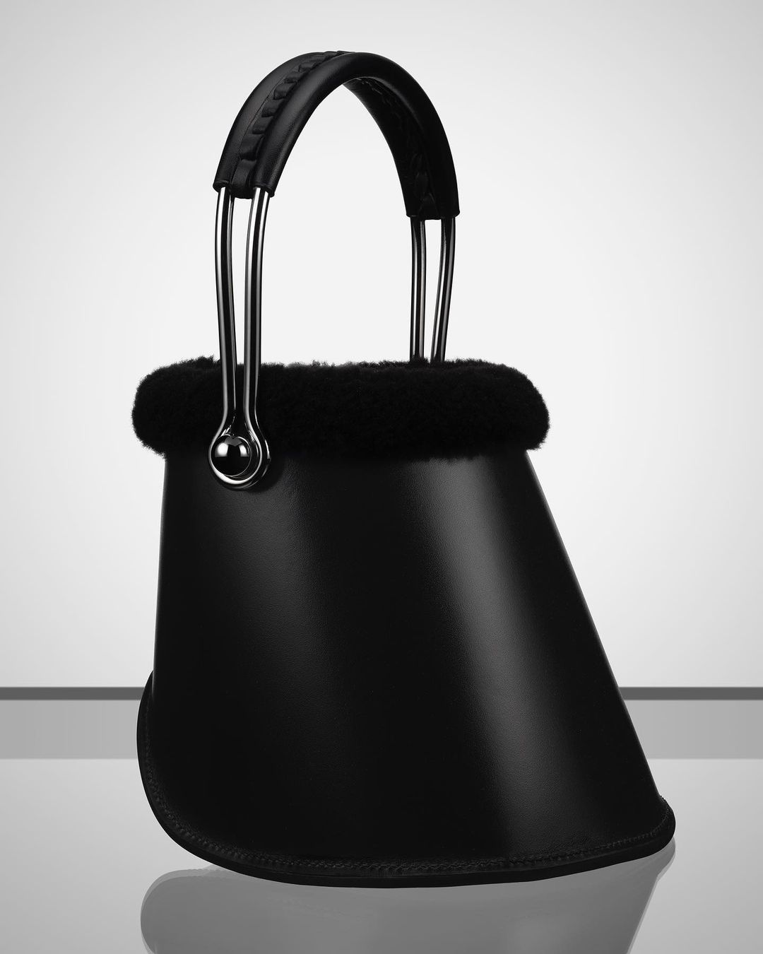 Studio shot of a black designer handbag with metal accents and a fur trim, set against a gradated grey and white background for fashion.