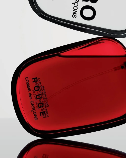 Product shot of a red Comme des Garçons Rouge perfume bottle, with black accents and text, positioned on a reflective surface.