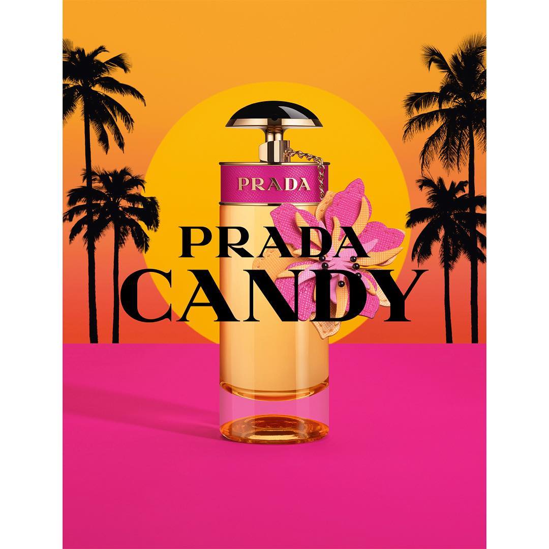 Prada Candy perfume bottle stands prominently against a vibrant backdrop of palm trees and a sunset-colored sky, creating a tropical feel.
