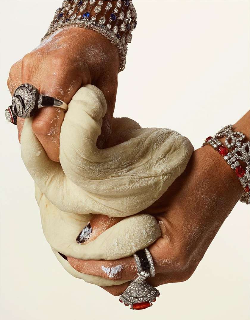 Close-up of two hands kneading dough, adorned with diamond bracelets and rings, against a plain white background. Food preparation, jewelry.