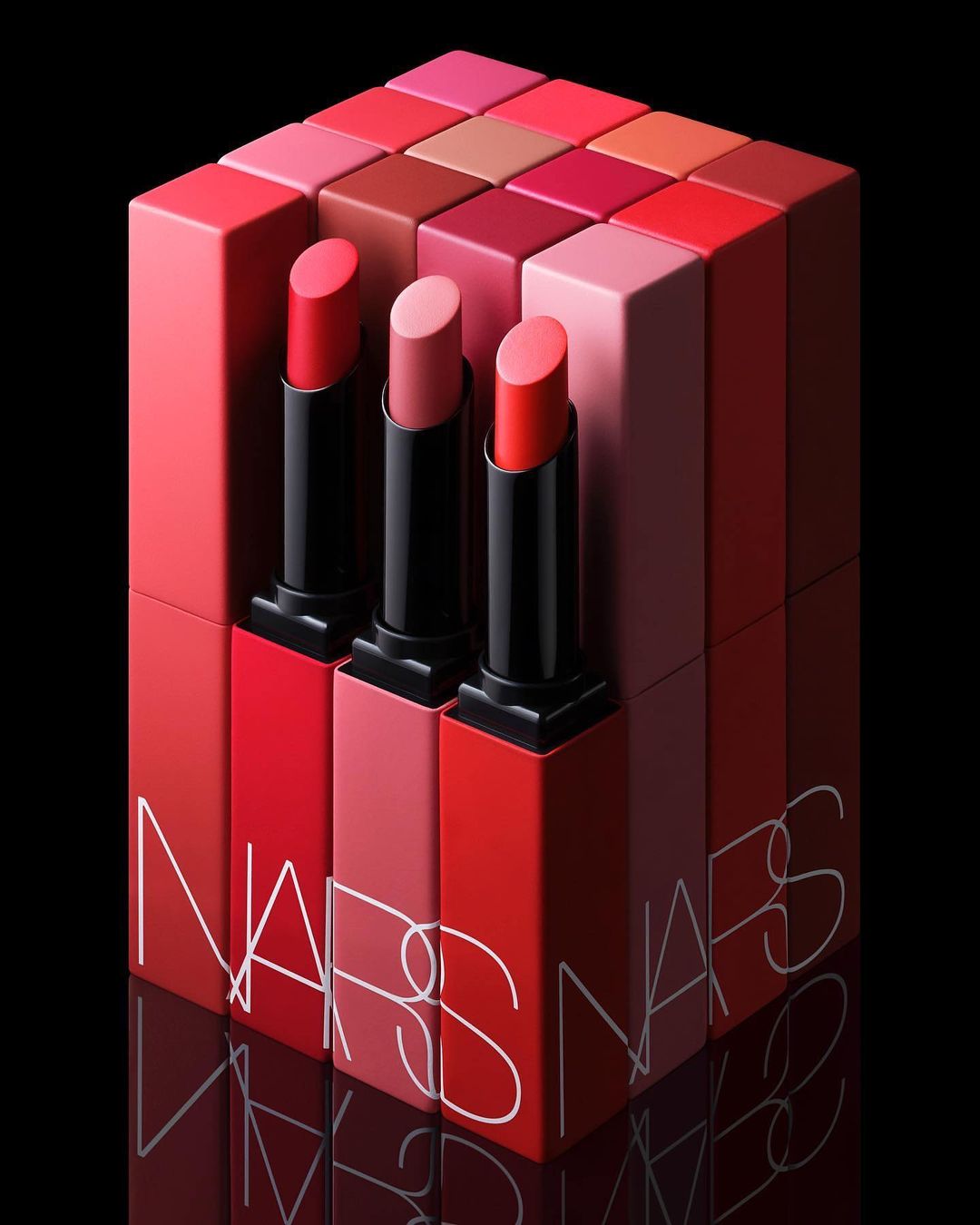 Three NARS lipsticks of varying shades stand in front of a display of red, pink, and brown lipstick boxes against a black background.