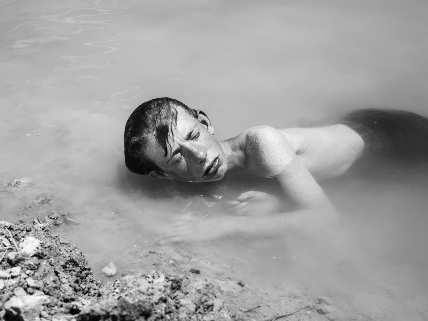 Black and white portrait of a young man lying in murky water, his eyes closed and his hair wet, with muddy banks nearby.