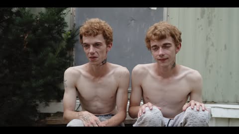 Two red-haired adult males with visible tattoos sit outside. They are wearing sweatpants, and green foliage is in the background.