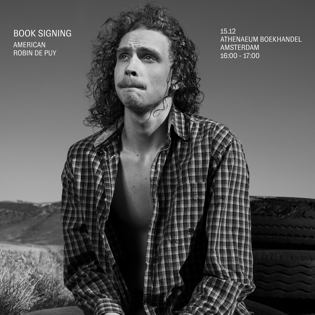 Black and white portrait of a man with curly hair and an open flannel shirt, sitting outdoors with a sky and tire background.