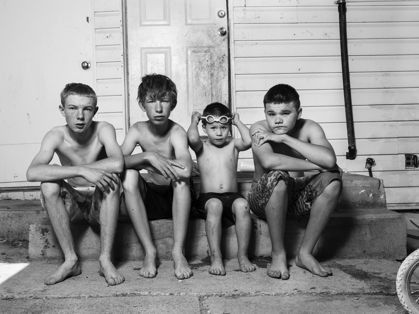 A black and white portrait of four boys sitting on a porch step with a closed door in the background, looking directly at the camera.