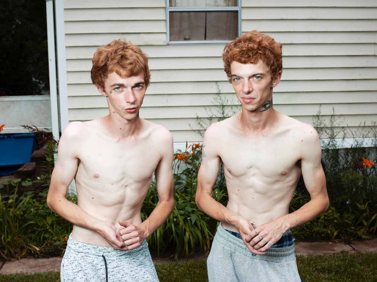 Outdoor portrait of two thin, red-haired adult males with light skin wearing grey sweatpants posing in front of a light-colored house and grass.