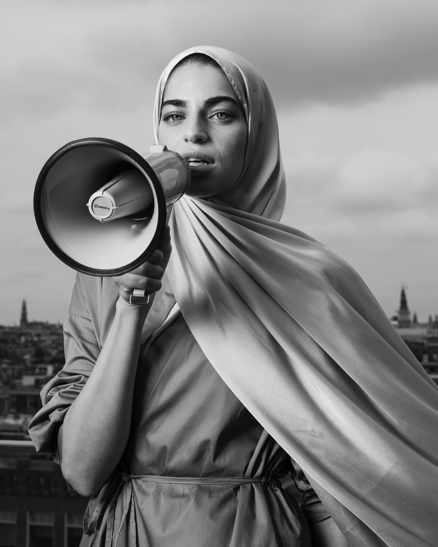 Black and white portrait of a woman in a headscarf speaking into a megaphone with a cityscape and cloudy sky in the background.