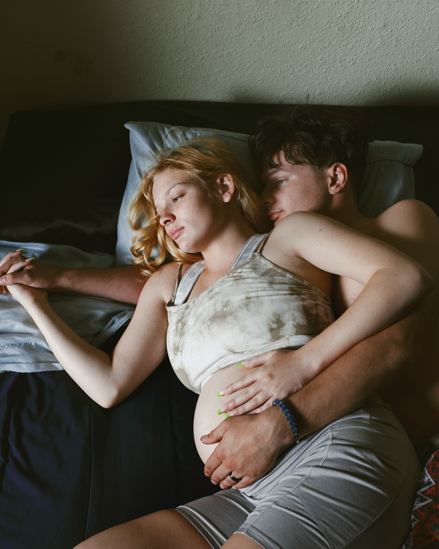 Pregnant woman and man embrace in bed, sharing an intimate moment in a softly lit room, showcasing a close-up perspective.