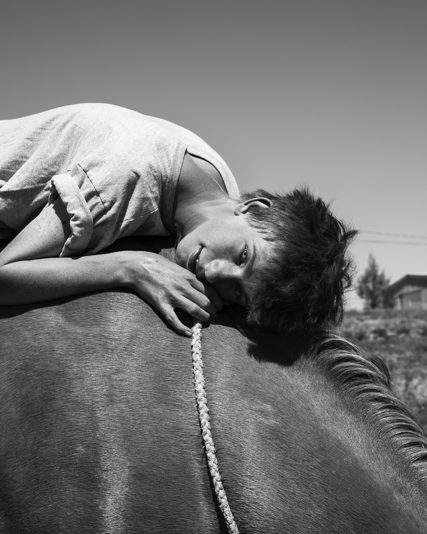 Black and white photograph of a woman lying on a horse's back outside on a sunny day with a clear sky, looking at the camera.