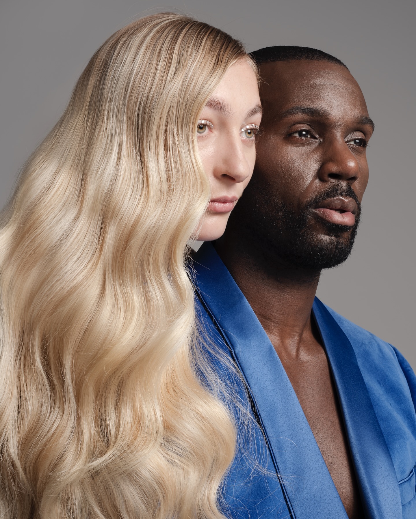Portrait of an adult Black man and adult Caucasian woman with wavy blonde hair, both looking off-camera in a neutral studio.