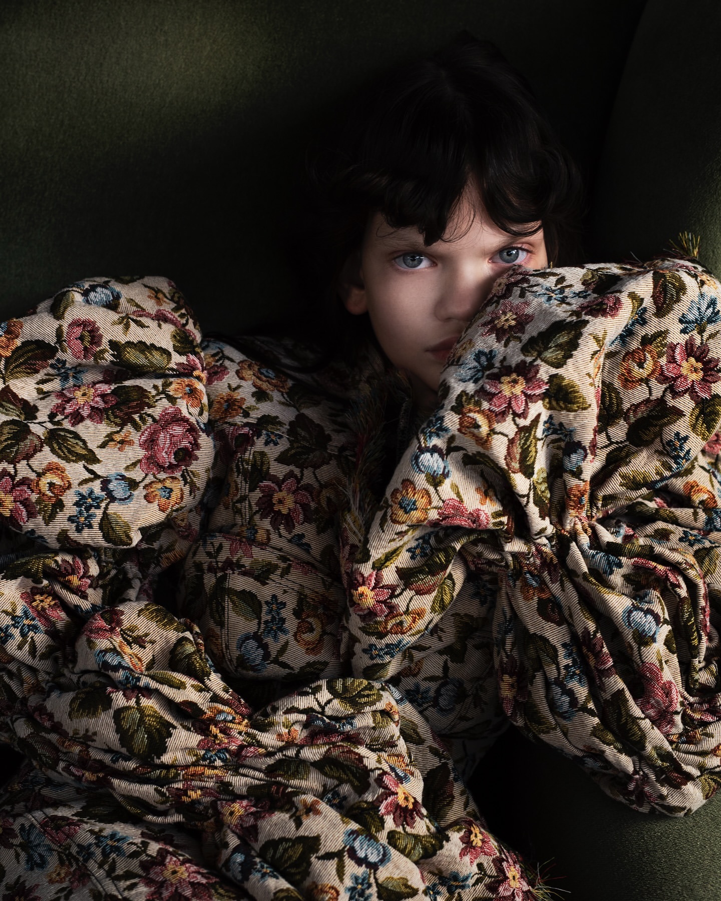 Child with dark hair and blue eyes is wrapped in a floral blanket on a green sofa, looking directly at the camera in a close-up shot.