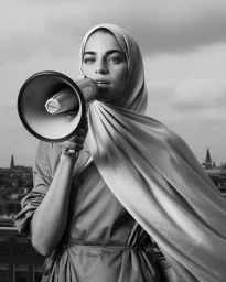 Black and white portrait of a woman wearing a hijab holding a megaphone up to her mouth against an outdoor skyline backdrop.