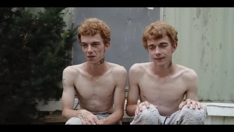 Portrait of two shirtless young twin men with red hair seated outdoors by a green bush and weathered building exterior.