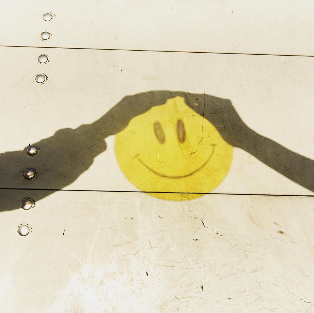 Yellow smiley face with black shadows suggesting hands holding it against a textured white surface, creating a playful and surreal image.