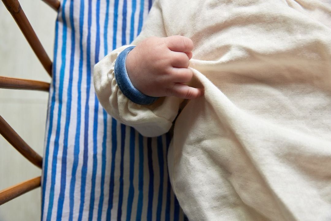 Close-up of a baby's hand gently holding a white cloth in a wooden crib with a blue and white striped patterned cushion.