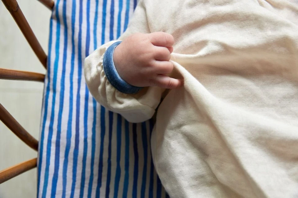 Close-up of a baby's hand gently holding a white cloth in a wooden crib with a blue and white striped patterned cushion.