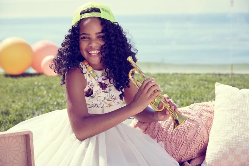 Smiling girl in a dress and neon cap sits on grass playing a gold trumpet on a sunny day outdoors near the ocean.