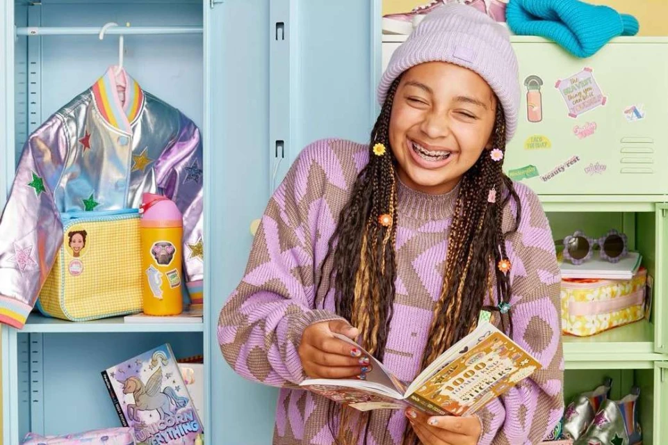 Smiling young woman with braces reads a sticker book in front of colorful lockers, wearing a purple knit hat and patterned sweater.