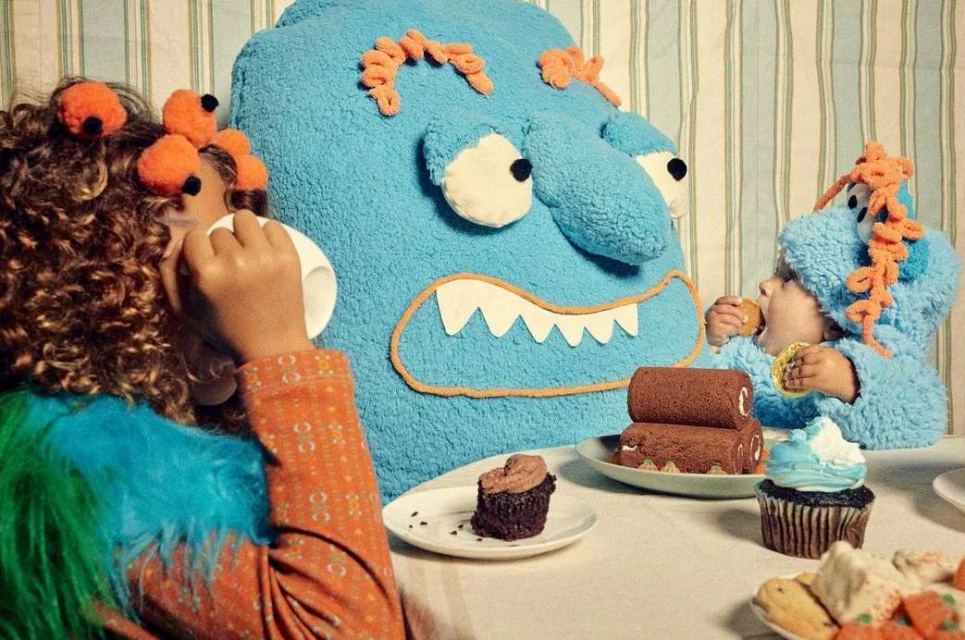 Child dressed in a monster costume eating dessert at a table with cupcakes, chocolate cake rolls, cookies, and a giant monster plush toy.