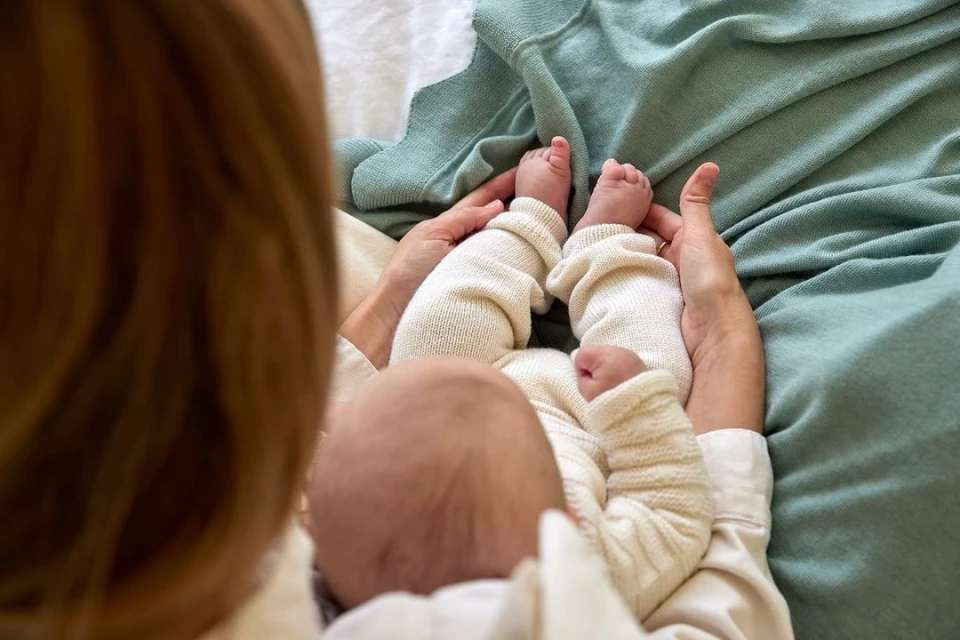A baby lying on their back with a woman's hands holding their legs and feet. The baby is wearing a white knit onesie.