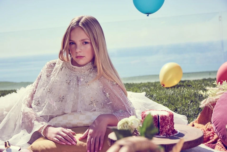 A young girl with long blonde hair poses in a formal dress with balloons and cake in an outdoor setting with an ocean view.