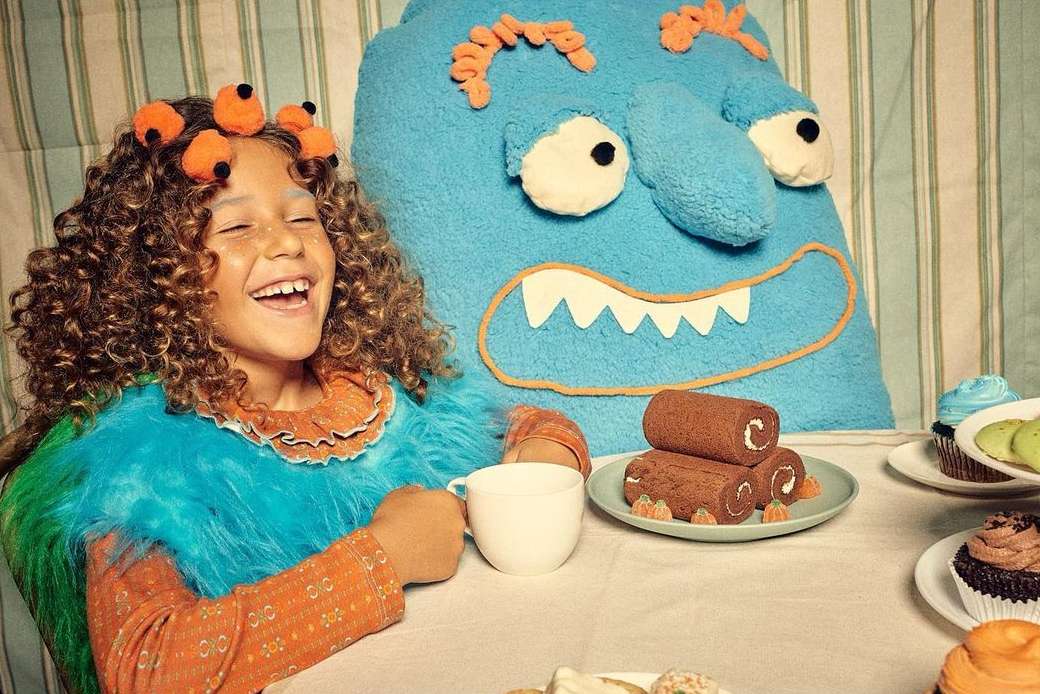 A young girl with curly hair laughs at a tea party with a blue monster plush, decorated with colorful sweets and pastries indoors.