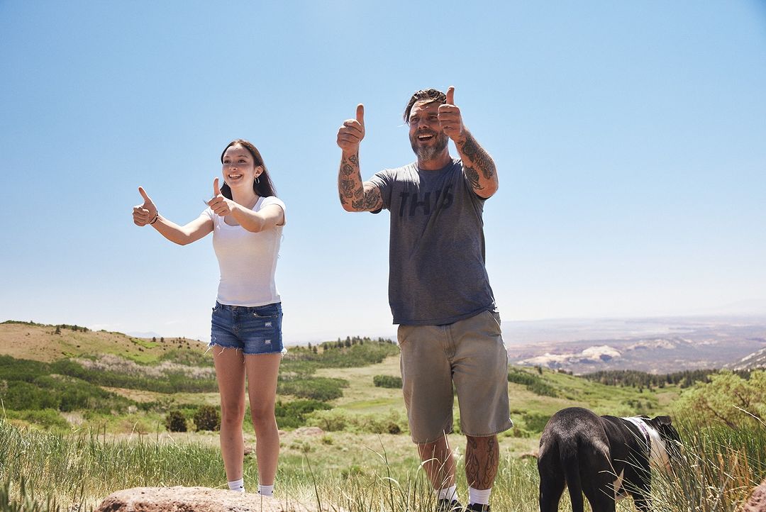 Father and daughter on a hilltop, smiling and giving thumbs-up gestures, with a dog standing nearby; clear blue sky, sunny outdoor scenery.