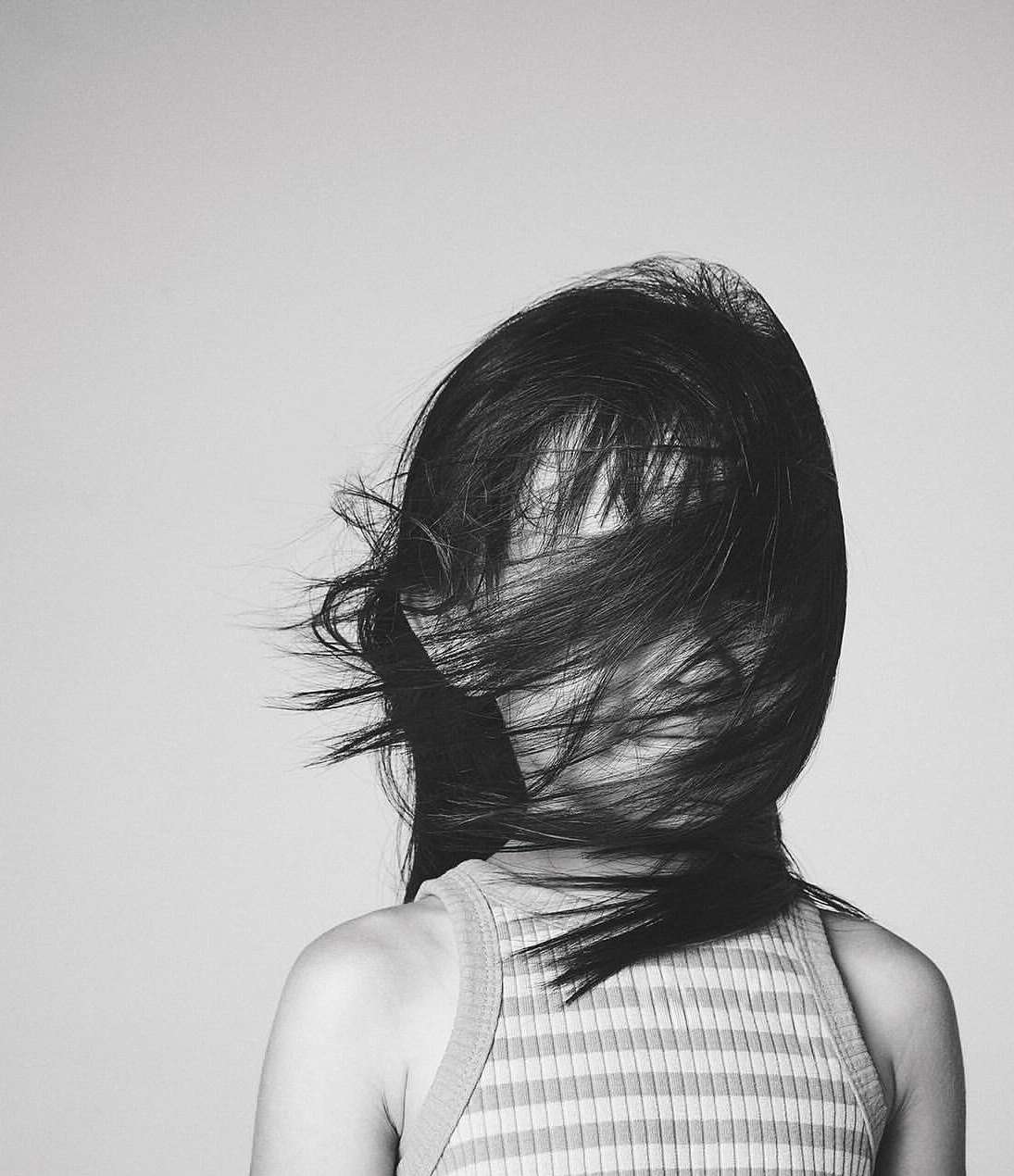 Monochromatic portrait of a woman with long hair partially obscuring her face, wearing a striped top against a light grey backdrop.