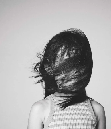 Monochromatic portrait of a woman with long hair partially obscuring her face, wearing a striped top against a light grey backdrop.