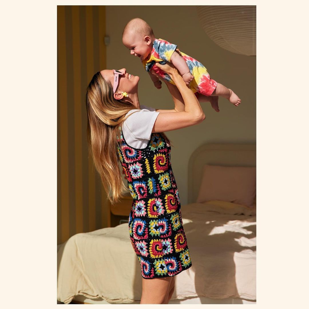 A woman holds a baby in the air, both smiling and wearing tie-dye clothing. The background shows a brightly lit interior room.